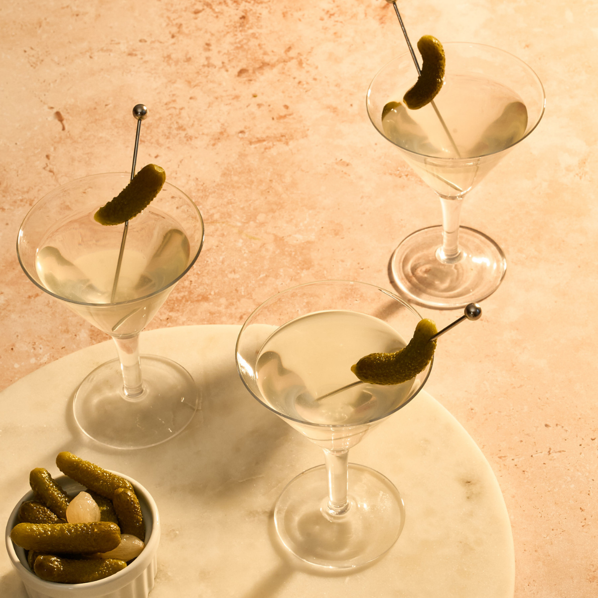 Three martini glasses with pickled green Cornichons on a marble tray against a pinkish-beige background.