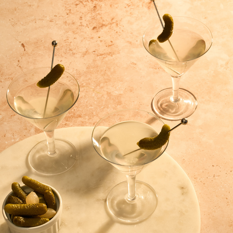 Three martini glasses with pickled green Cornichons on a marble tray against a pinkish-beige background.