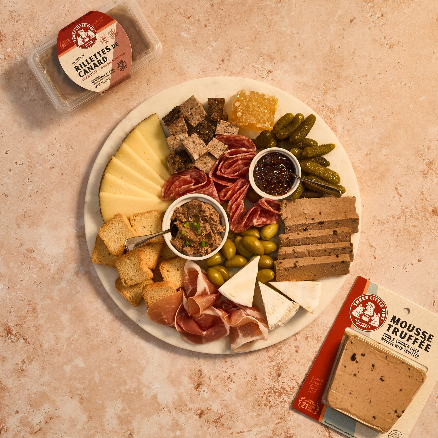 Platter of assorted meats, cheeses, and pickles with a small container of rillettes on a textured surface.