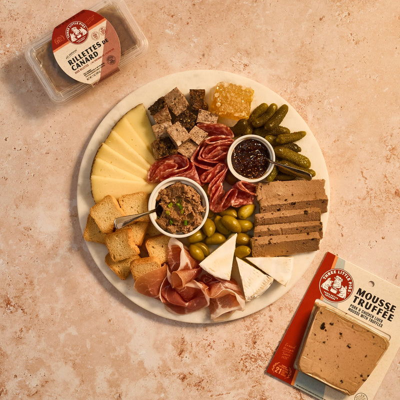 Platter of assorted meats, cheeses, and pickles with a small container of rillettes on a textured surface.