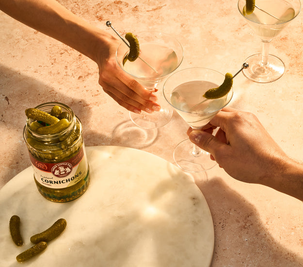 Two people holding cocktails with Cornichons near a jar of pickled cornichons on a marble surface.